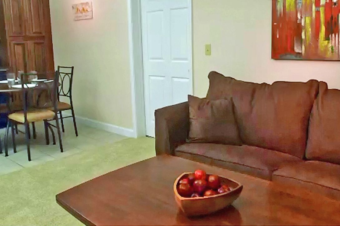 A living room with a brown leather sofa, a wooden coffee table with a bowl of fruit, a dining area with a wooden table and chairs, and a white door.