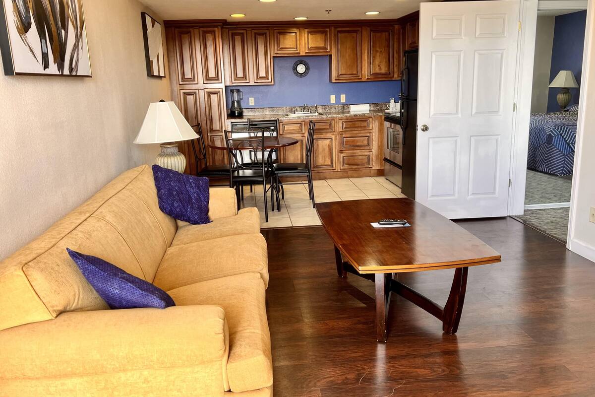 A living room with a sofa, a coffee table, and a dining area with chairs. There is a kitchen in the background with wooden cabinets and a refrigerator.