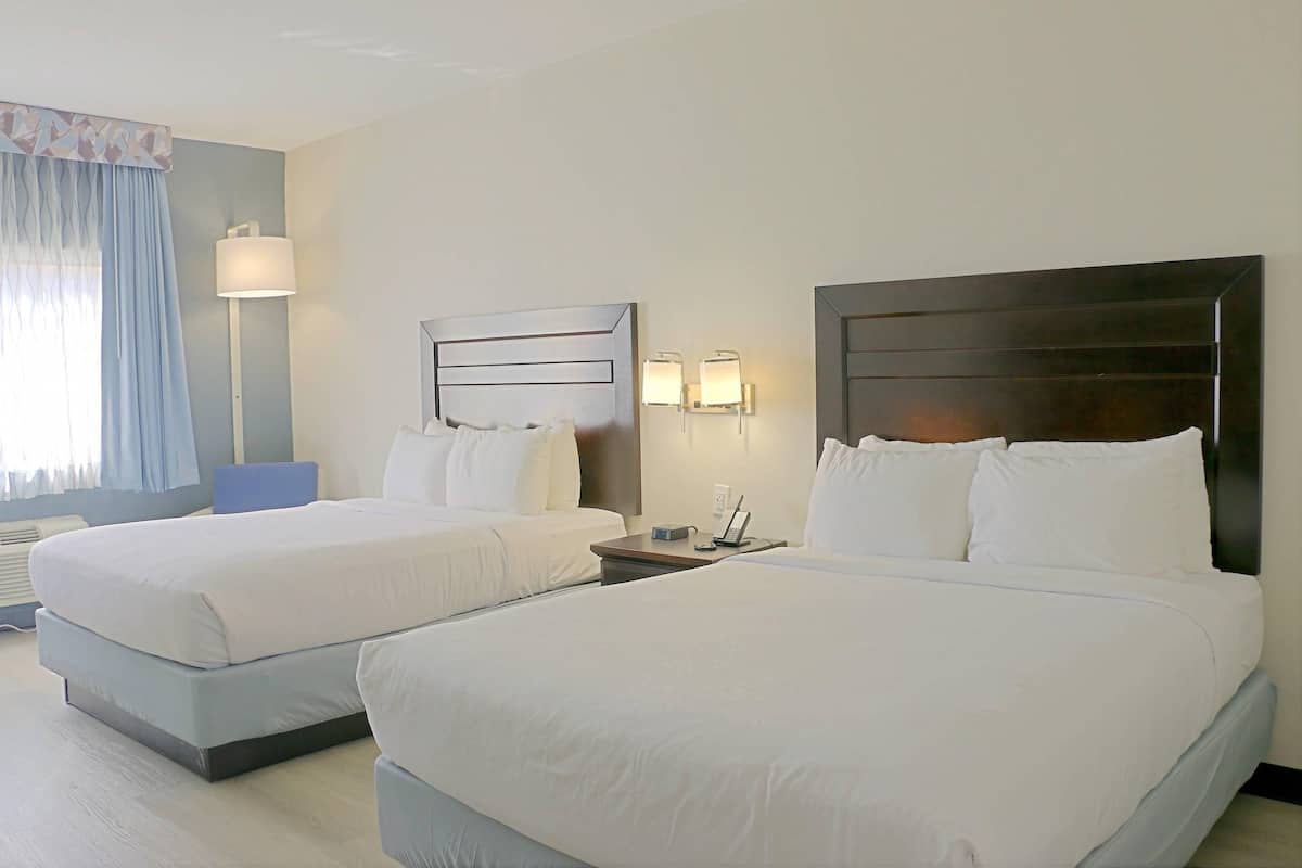 Quarto de hotel com duas camas, uma mesa de cabeceira com abajur e uma janela com cortinas.