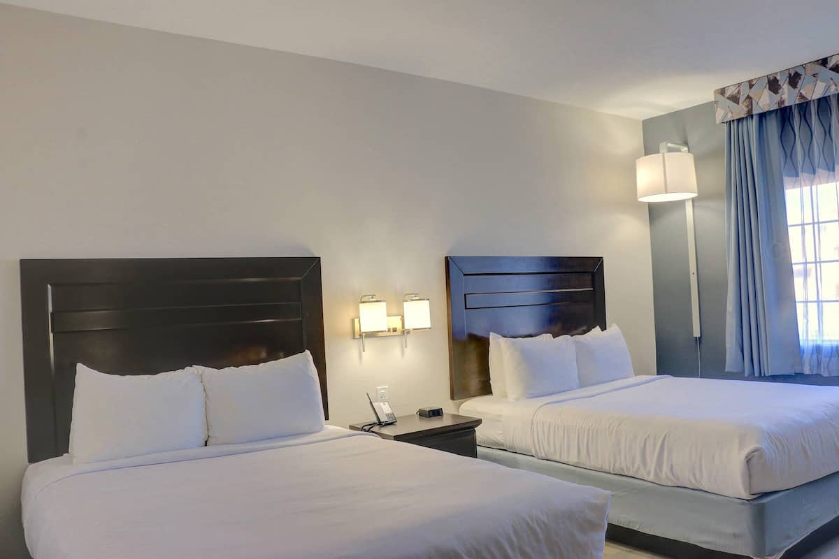 Quarto de hotel com duas camas, uma mesa de cabeceira com telefone, luminárias na parede e uma janela com cortinas.