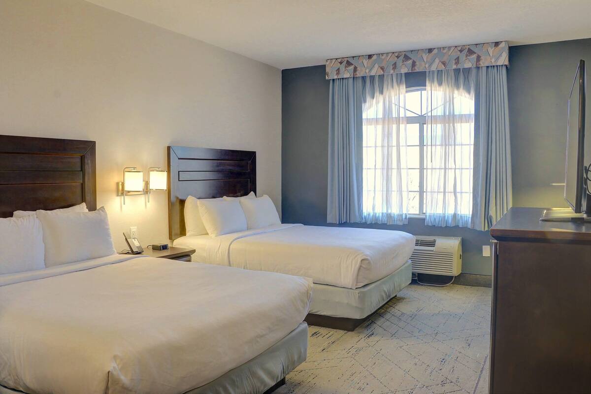 Quarto de hotel com duas camas, uma mesinha de cabeceira com telefone, escrivaninha com luminária e janela com cortinas.