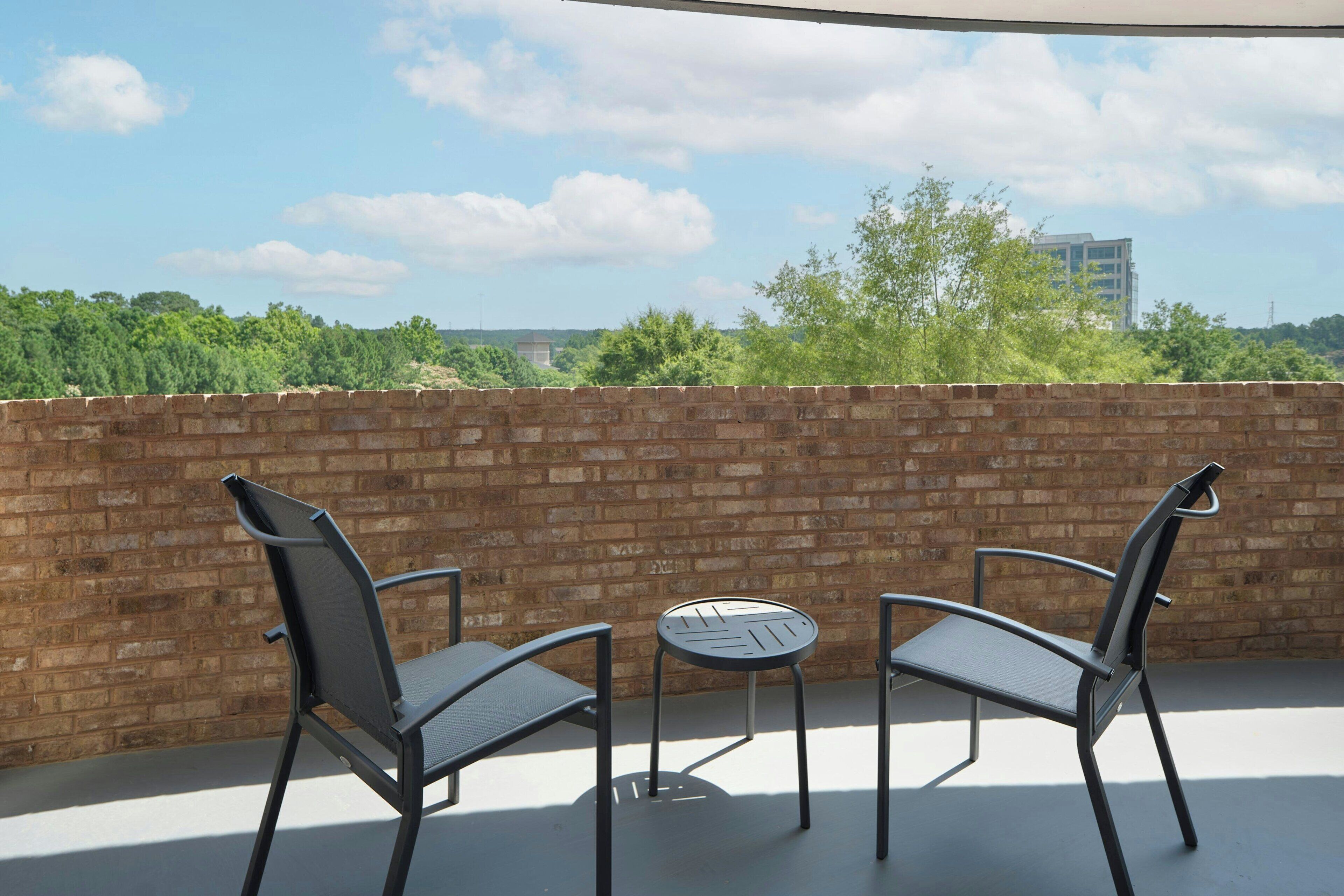A balcony with two chairs and a small round table, a brick wall, and a view of greenery and buildings in the distance.
