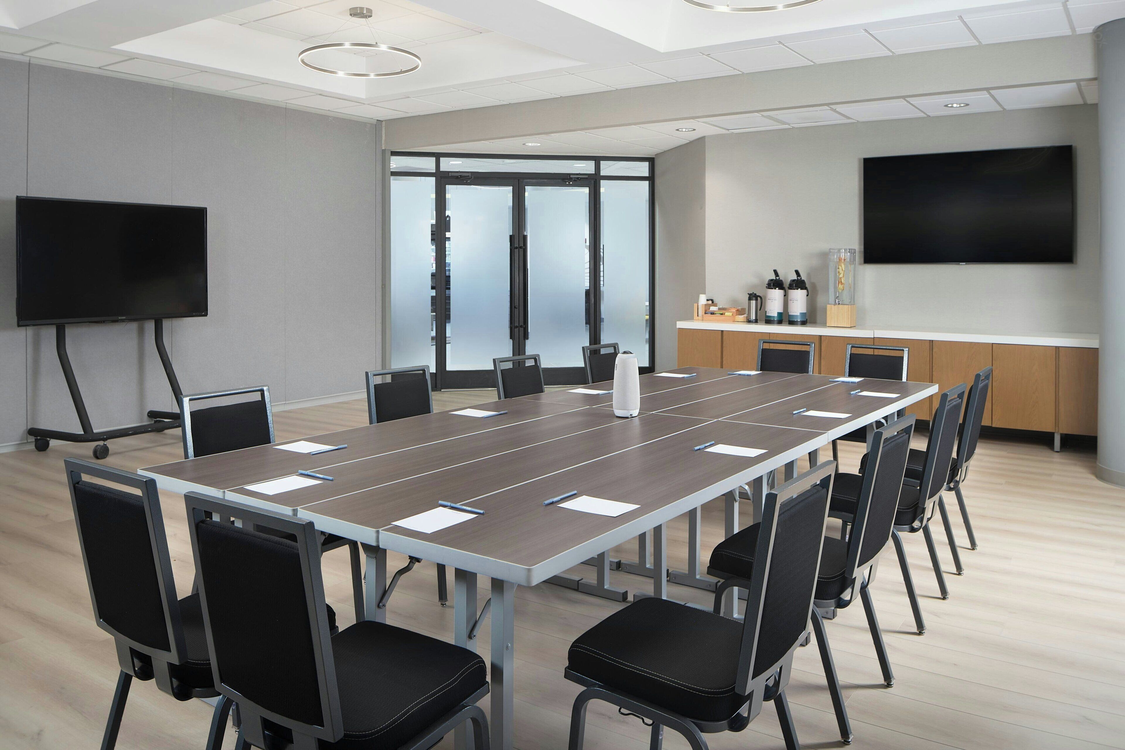 A conference room with a long table, chairs, a TV, and a glass door leading to a balcony.