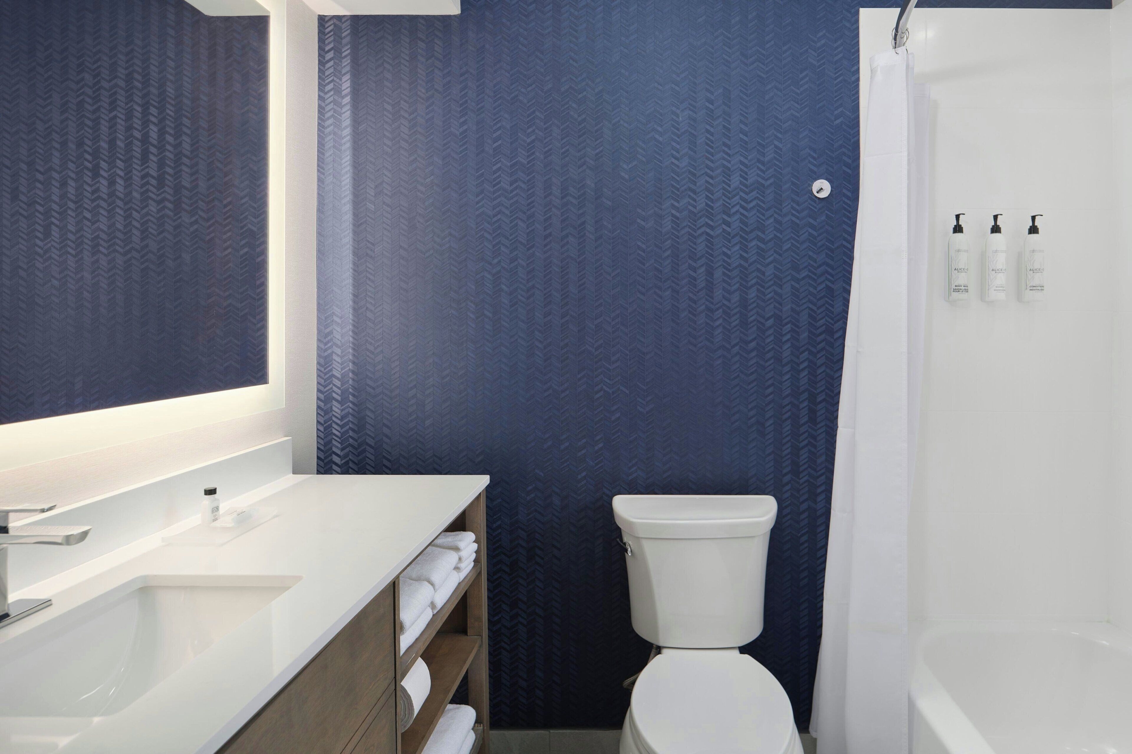 A modern bathroom with a white sink, mirror, and toilet. There are three bottles of toiletries on the wall.