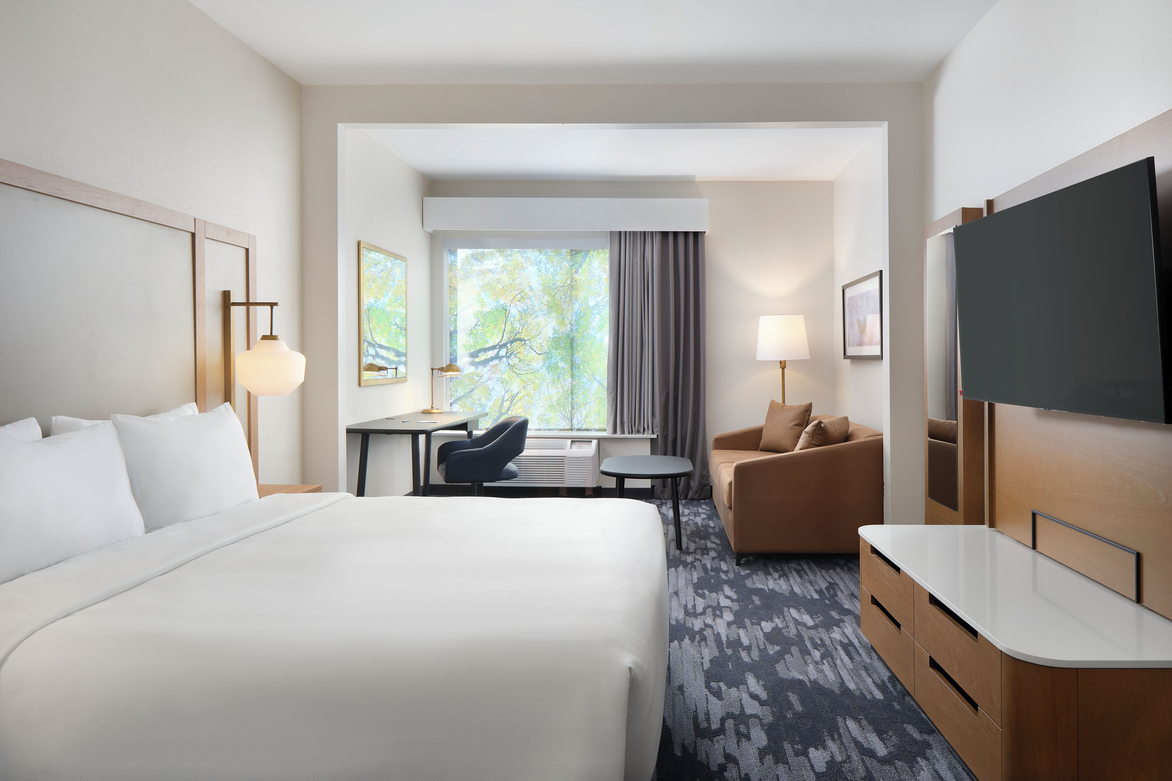 A hotel room with a large bed, a desk, a chair, a sofa, a TV, and a window with curtains.