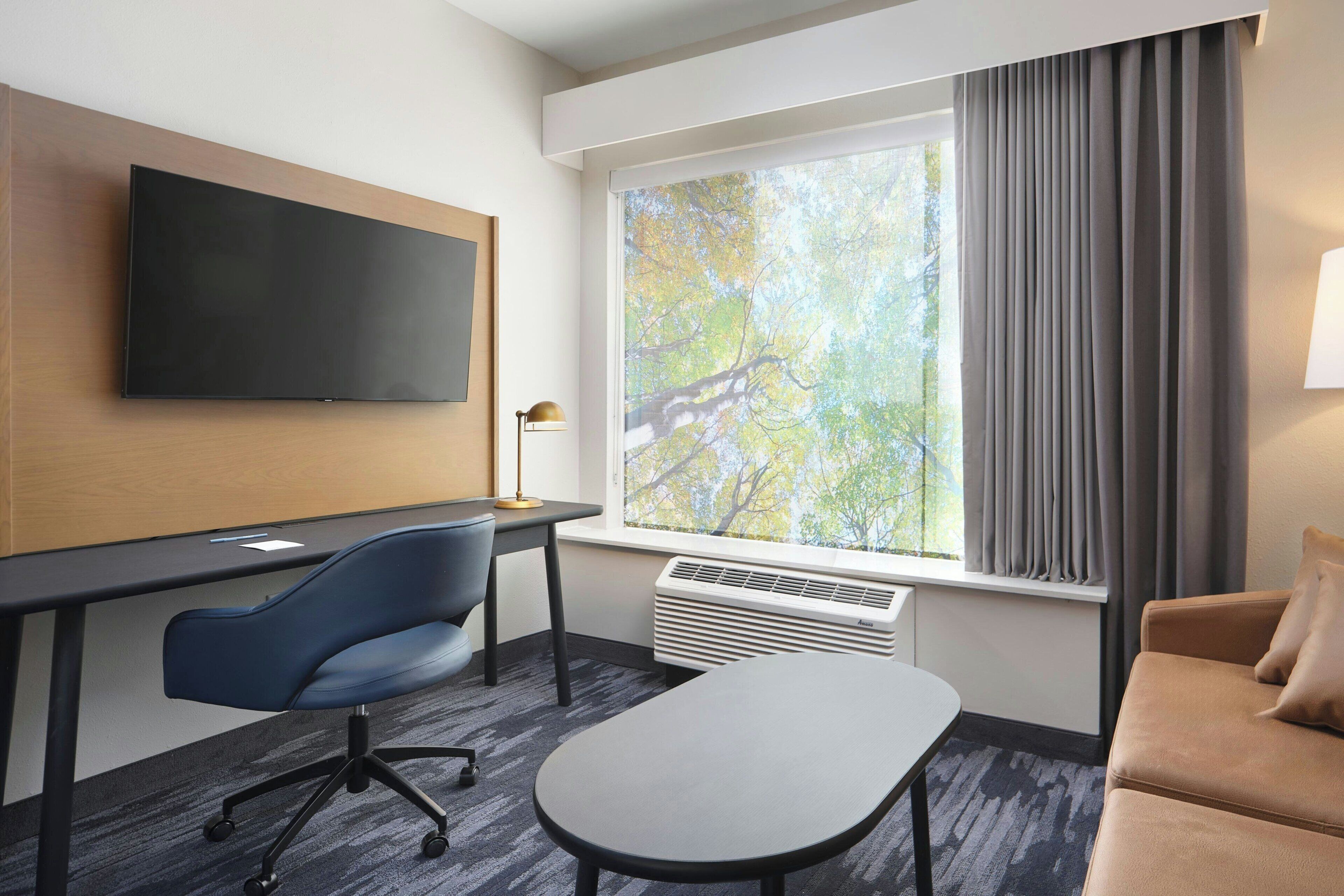 Une chambre d’hôtel moderne dotée d’un téléviseur à écran plat fixé au mur, d’une grande fenêtre avec des rideaux, d’un bureau avec une chaise, d’un canapé et d’un lampadaire.
