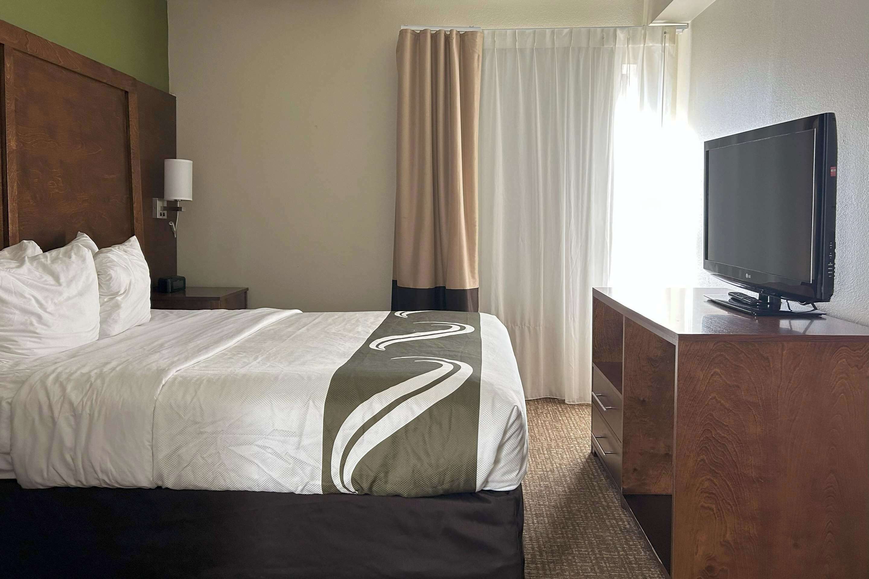 A hotel room with a bed, a desk with a television, a lamp, and a window with curtains.