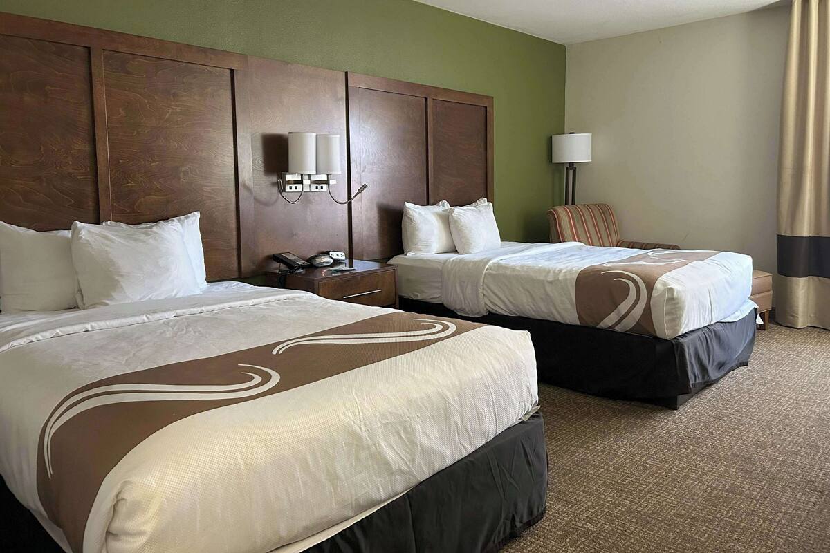 A hotel room with two beds, a wooden headboard, a nightstand with a phone, and a lamp.