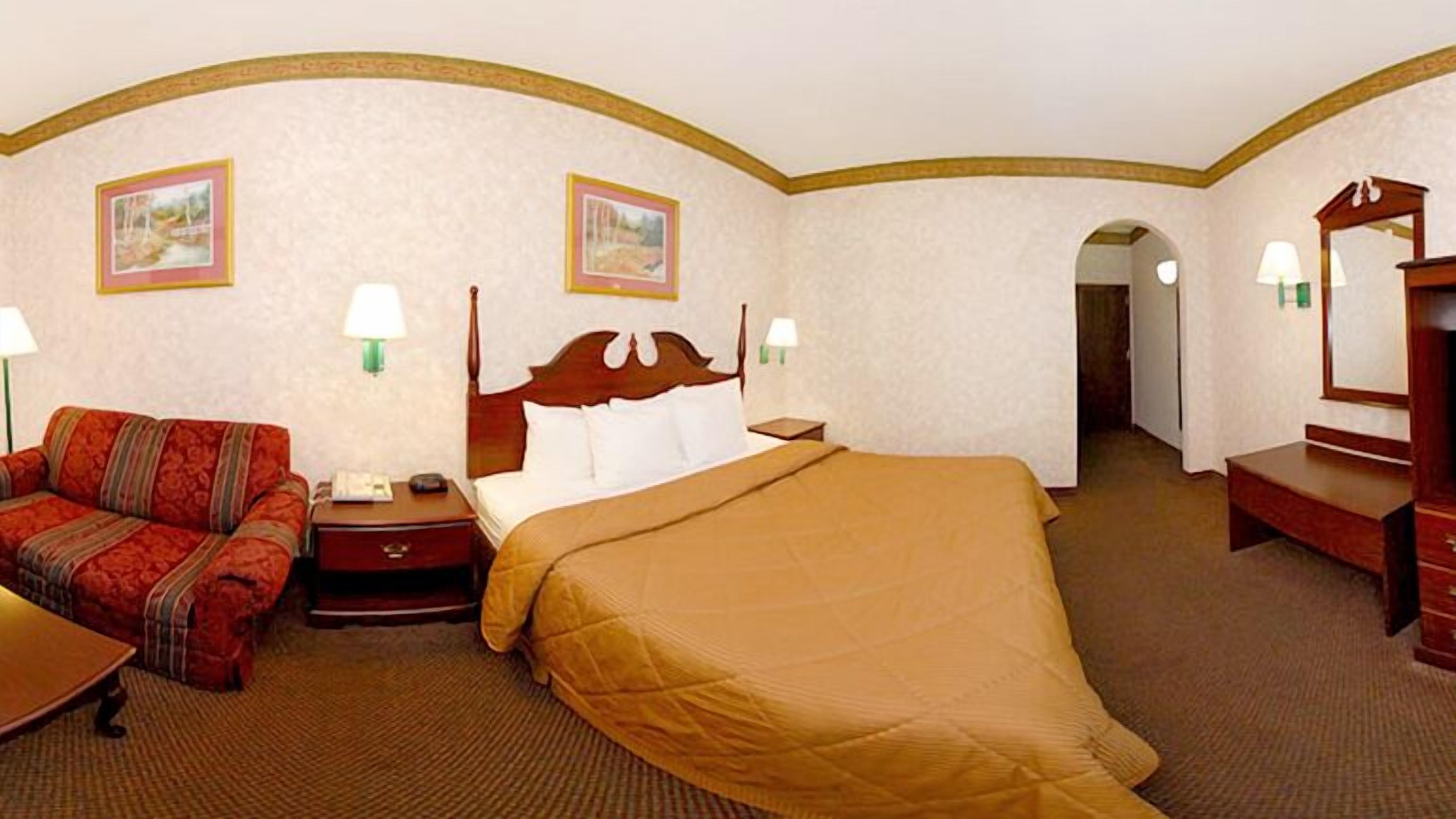 Una habitación de hotel con cama, sofá, mesita de noche, lámpara y espejo.