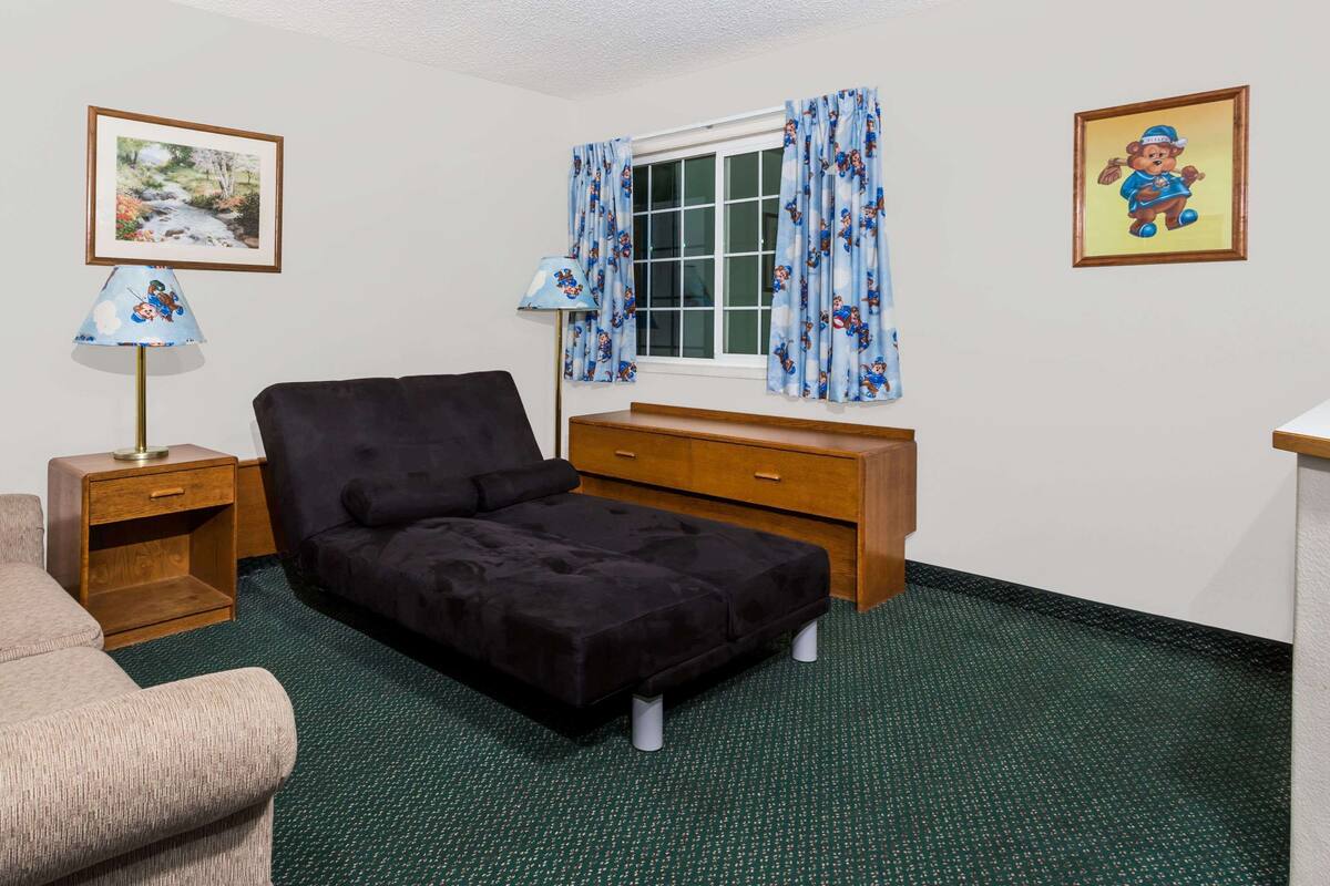 Una habitación con un sofá negro, una mesita de noche de madera, un pequeño aparador de madera y una ventana con cortinas azules.