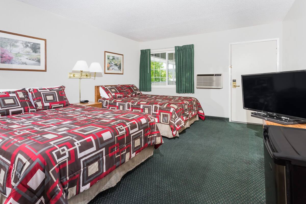Habitación de hotel con dos camas, televisión, ventana con cortinas, lámpara y un cuadro enmarcado en la pared.