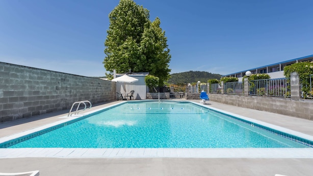 A rectangular swimming pool with clear blue water, surrounded by a concrete deck. There are white lounge chairs and a small covered seating area with a table and chairs. A tall tree is visible behind the pool, and a building with a blue roof and white walls is in the background.