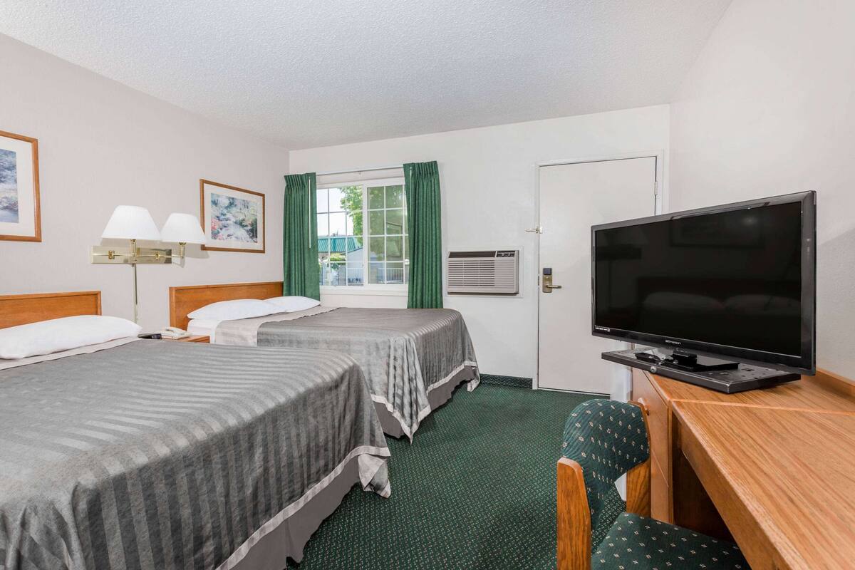 Habitación de hotel con dos camas, un escritorio con televisor, una silla, una lámpara y una ventana con cortinas.