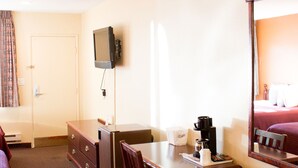 Quarto de hotel com cama, escrivaninha com cadeira, televisão presa à parede e espelho sobre a escrivaninha.