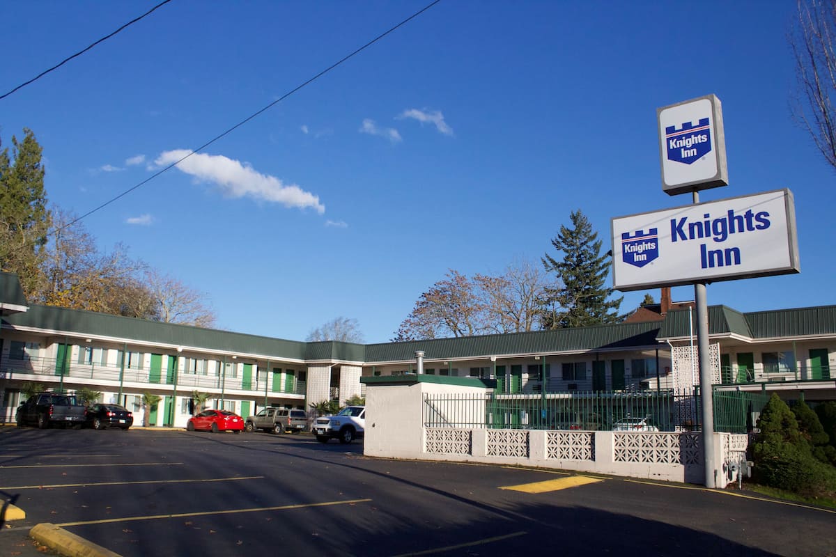 Un motel Knights Inn à l’extérieur vert et blanc, doté d’un parking clôturé et surplombant un ciel bleu dégagé.