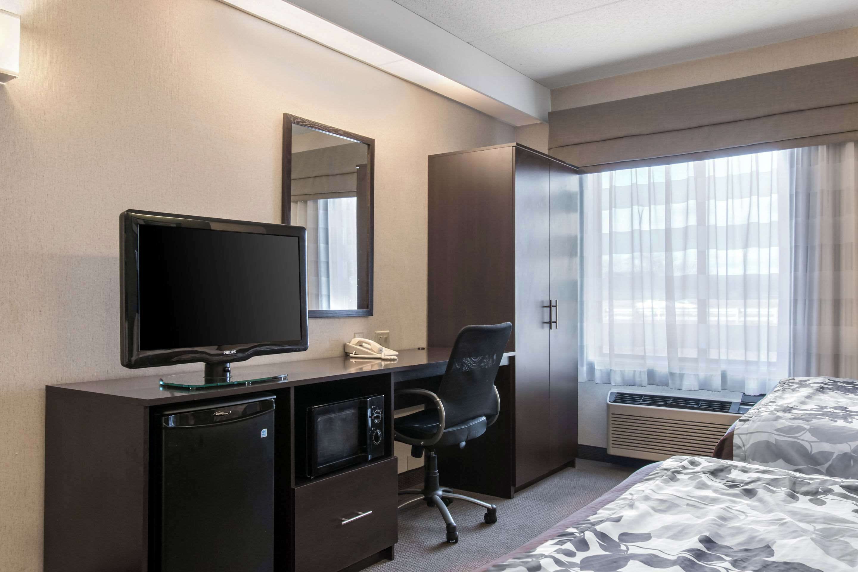 A hotel room with a flat-screen TV, a desk with a phone, a black office chair, a mini fridge, a bed with a camouflage-patterned bedspread, and a large window with sheer curtains.