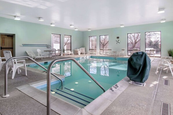A swimming pool with clear lanes, surrounded by white chairs and tables, and large windows offering views of trees.