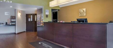 A hotel reception area with a wooden reception desk, a coffee machine, and a 'Welcome' mat.