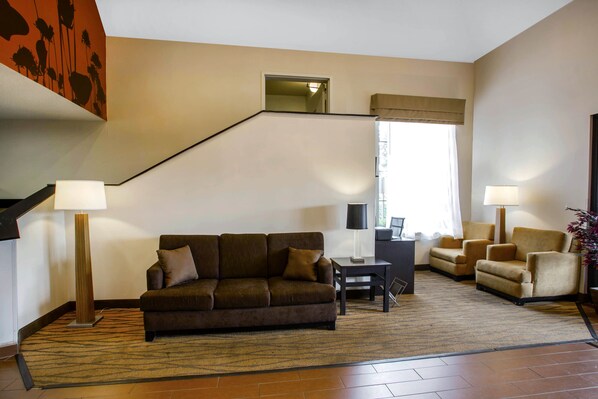 A living room with a brown sofa, two armchairs, a coffee table, and a rug. There is a staircase with a mirror and a window with curtains. - Sleep Inn Nashville - Brentwood - Cool Springs (Brentwood)