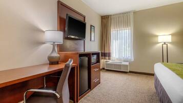 A hotel room with a wooden desk, a chair, a television, a bed, a window with curtains, and a lamp.