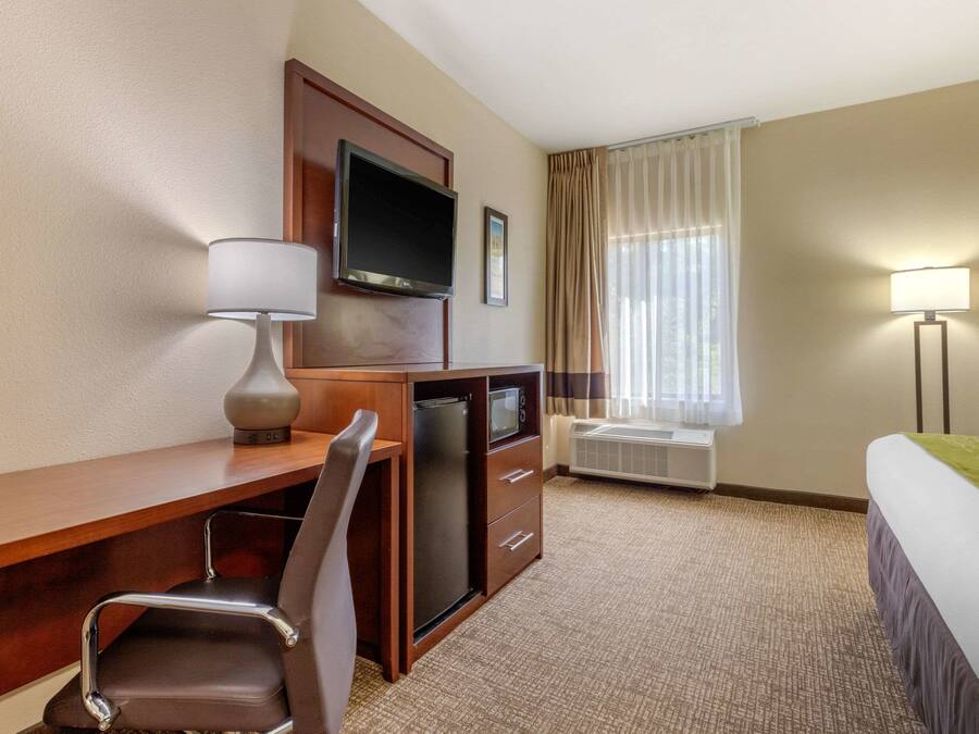 A hotel room with a wooden desk, a chair, a television, a bed, a window with curtains, and a lamp.