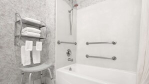 Un baño con una bañera blanca, una ducha con cabezal de ducha manual, un estante de metal con toallas y un taburete.