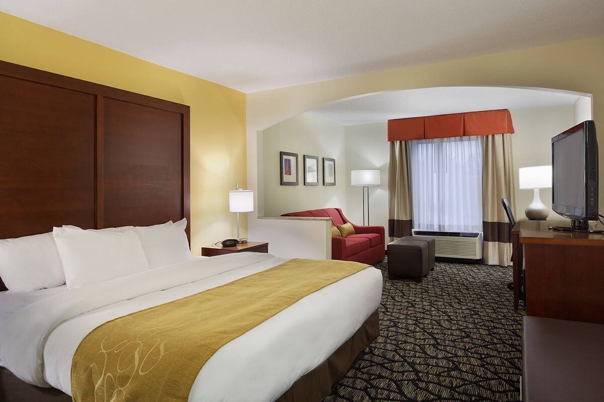 A hotel room with a large bed, a desk with a television, a red armchair, and a small table with a lamp.