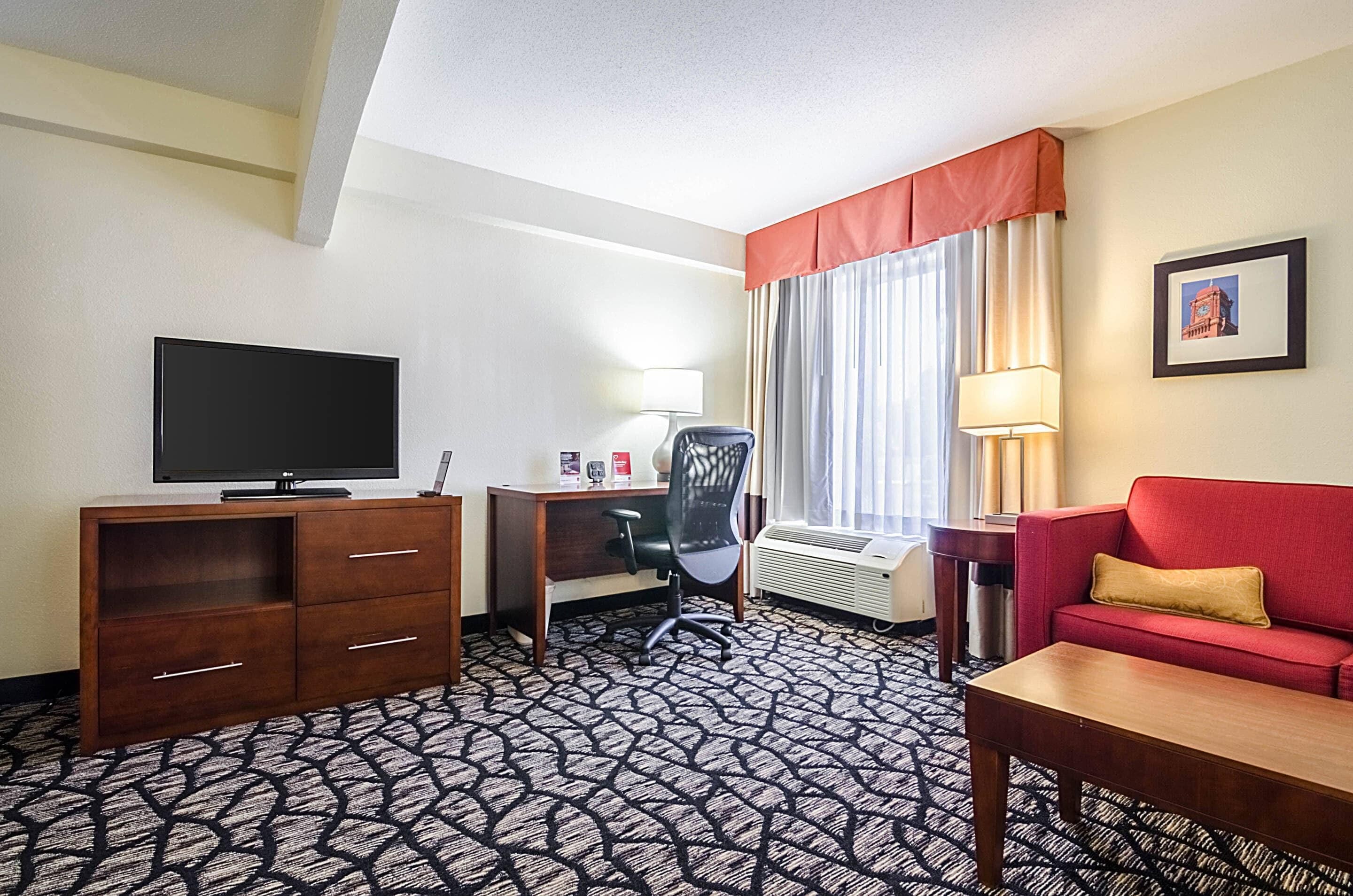 A hotel room with a flat-screen TV, a desk with a chair, a red sofa, and a patterned carpet.