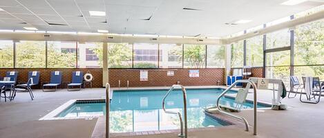 An indoor swimming pool with clear glass walls, blue lounge chairs, and a brick wall with windows.