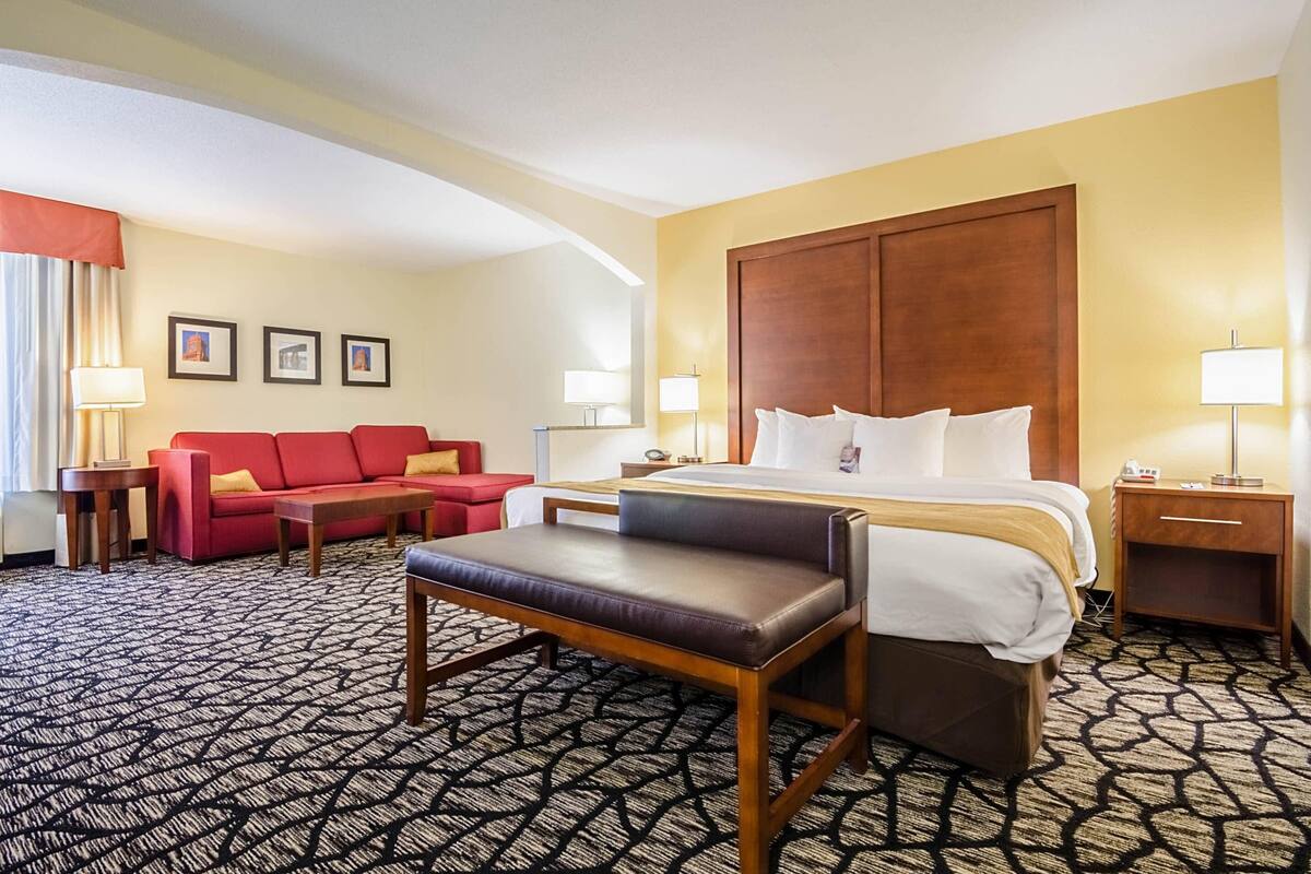 A hotel room with a bed, a red sofa, a brown chair, a small table, and a nightstand with a lamp.