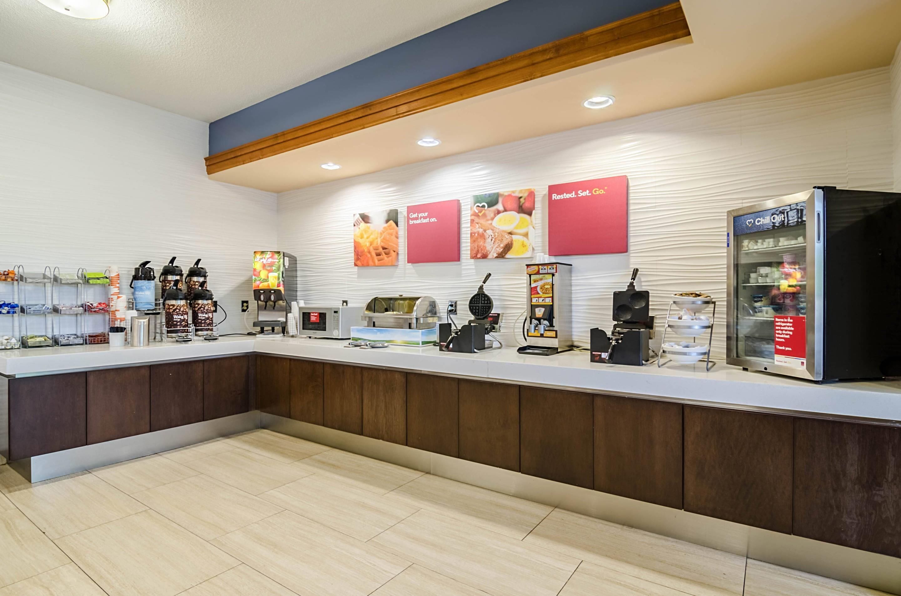A hotel reception area with a counter, coffee machines, a refrigerator, and promotional posters on the wall.
