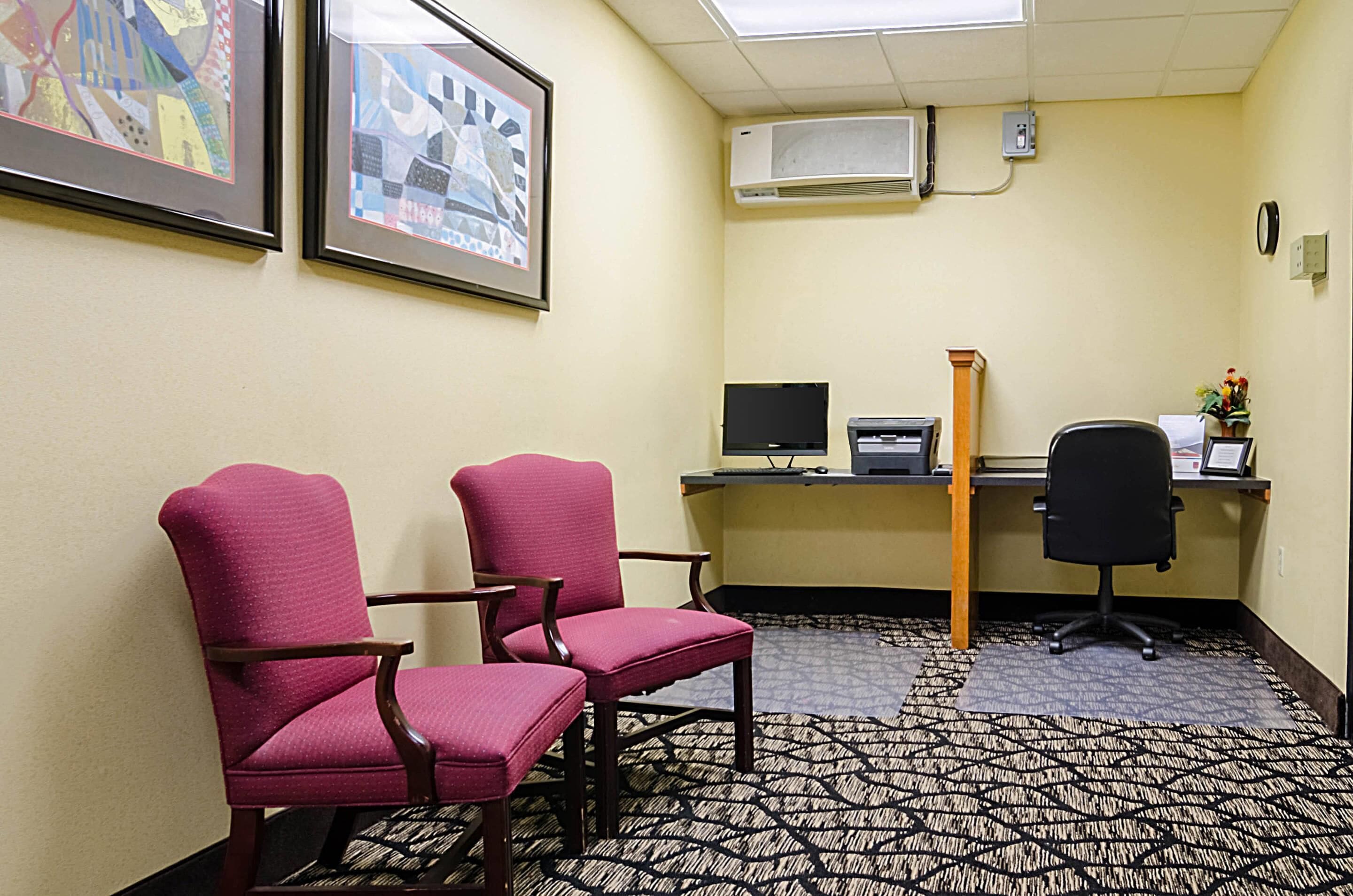 A hotel room with two pink chairs, a desk with a computer, and an air conditioning unit on the wall.