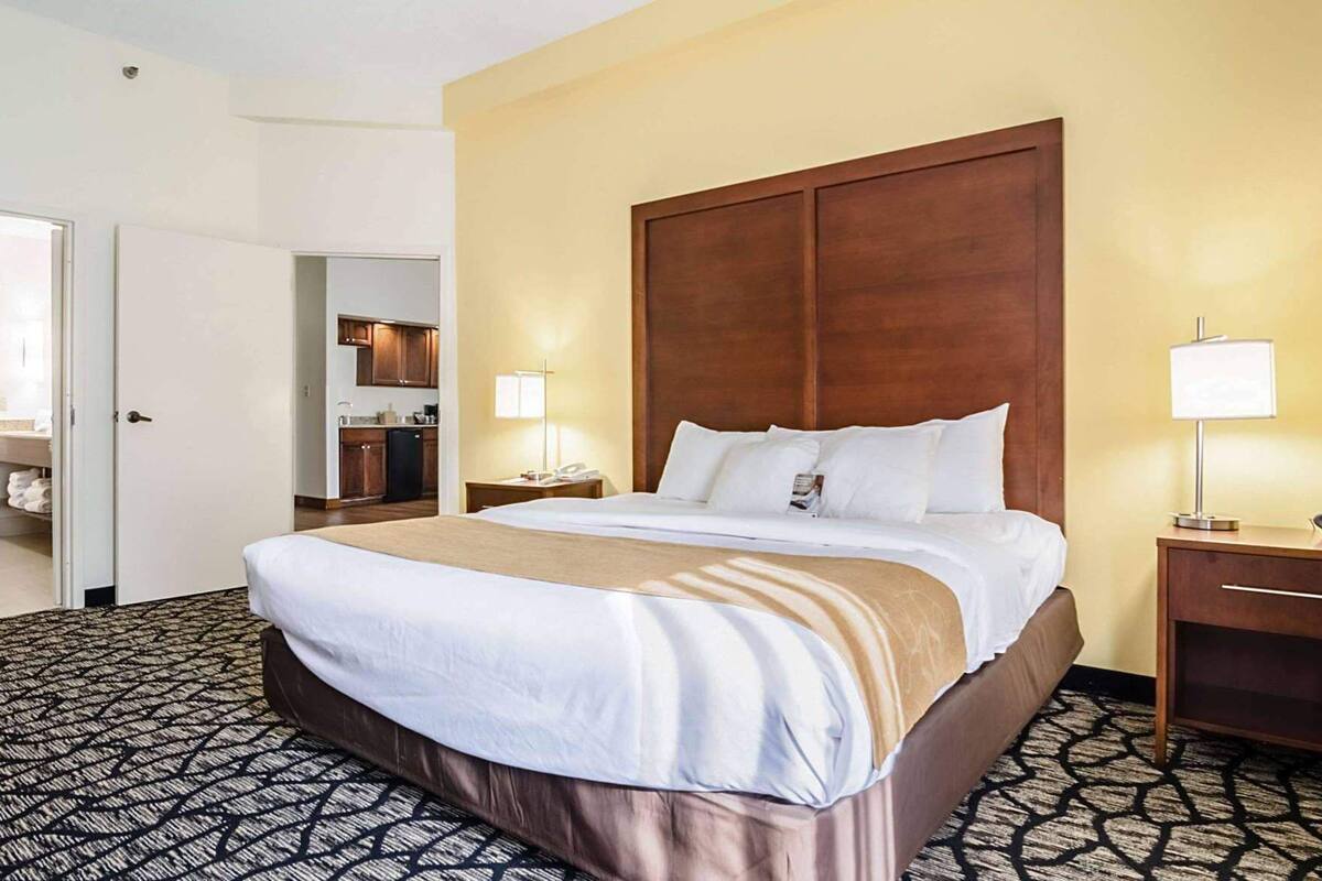 A hotel room with a large bed, a wooden headboard, a bedside table with a lamp, and a small desk with a telephone.