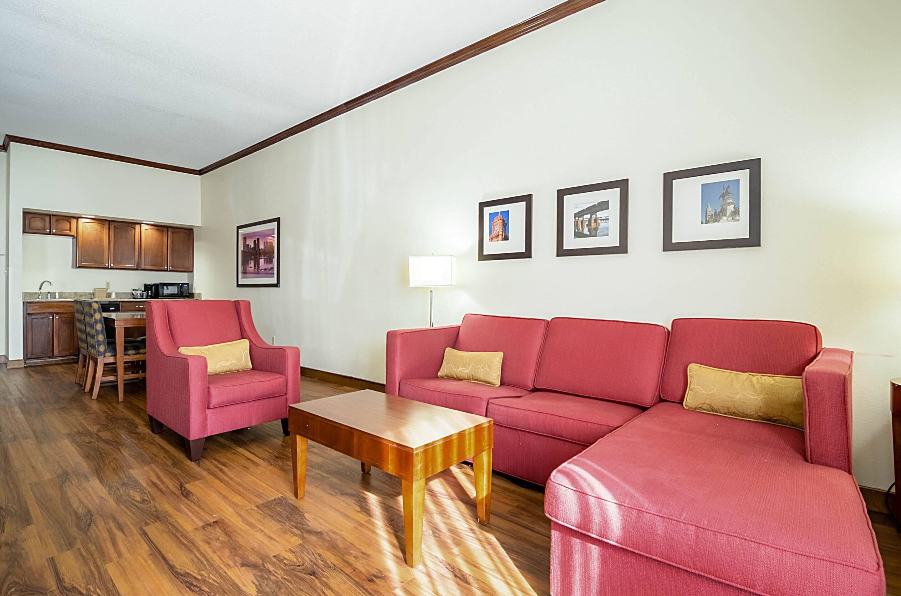 A living room with a red sofa, a wooden coffee table, a red armchair, a dining table with chairs, and a kitchen in the background.