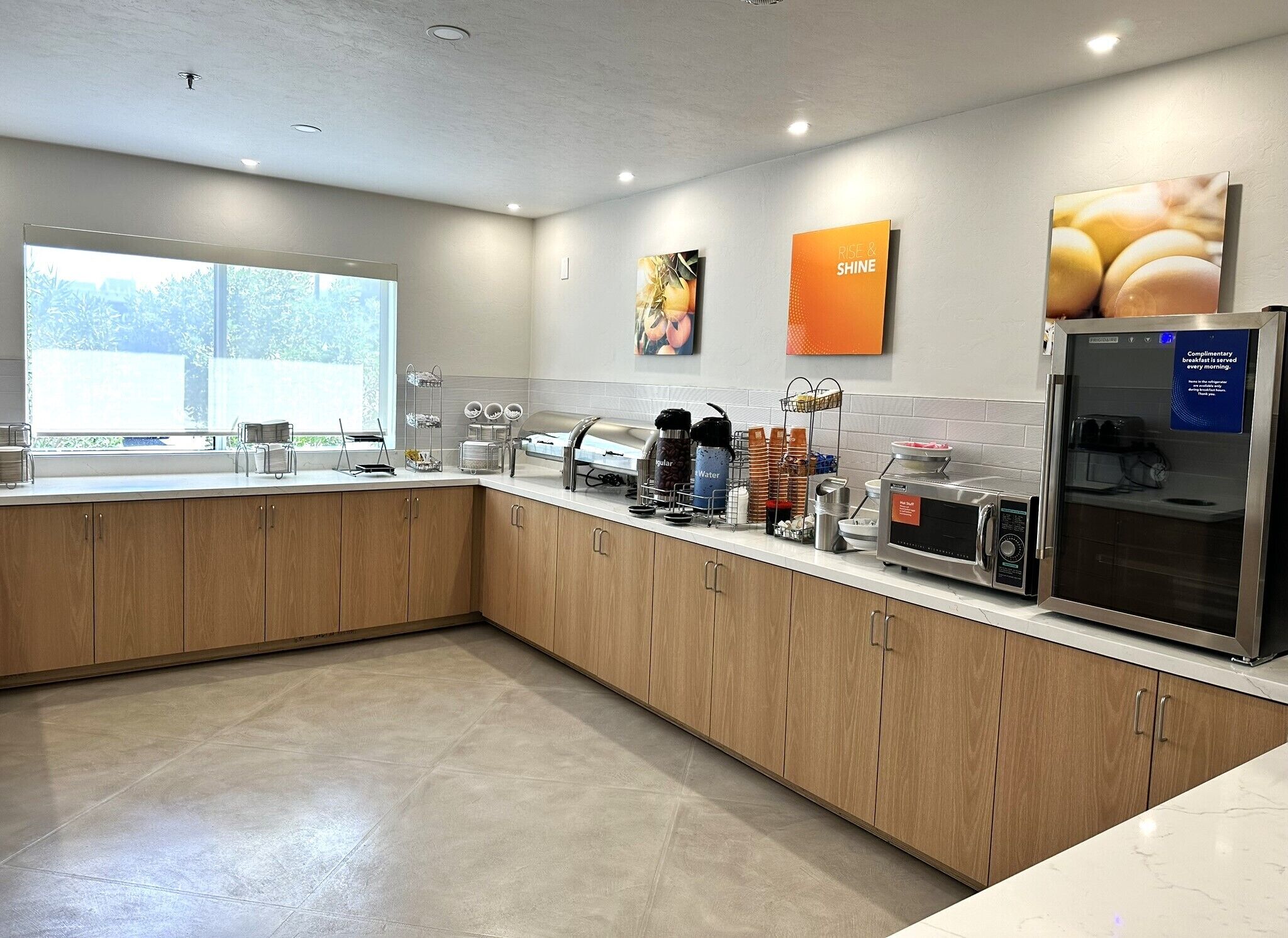 A modern kitchen with wooden cabinets, a stainless steel refrigerator, a microwave, and various kitchen appliances. There are two windows providing natural light, and a large window on the left side.