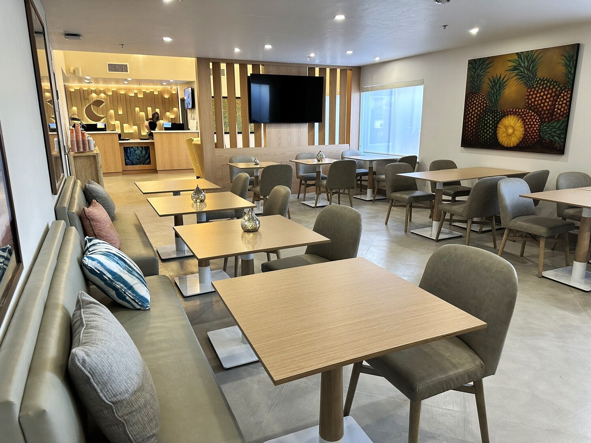 A modern restaurant with wooden tables and cushioned seating, a bar area, and a large pineapple artwork on the wall.