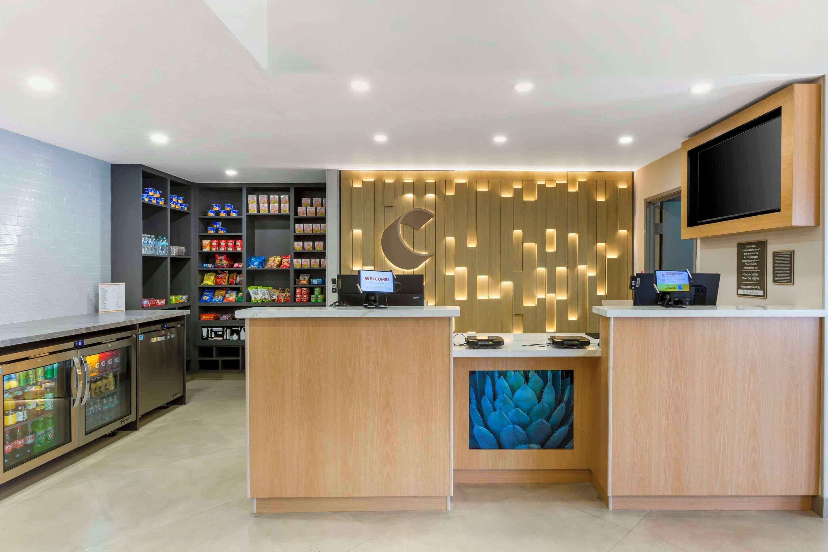 A reception area with a wooden reception desk, a digital screen, and a wall-mounted TV. There are shelves with snacks and beverages, and a refrigerator with a glass door.