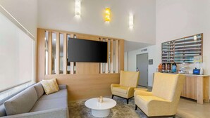 A modern hotel lobby with a grey sofa, two yellow armchairs, a white coffee table, a flat-screen TV mounted on a wooden panel, and a bar area with various amenities. - Comfort Suites At Tucson Mall (Tucson)