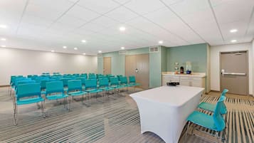 Uma sala de conferências com fileiras de cadeiras azul-turquesa, uma mesa branca e uma área de cozinha.