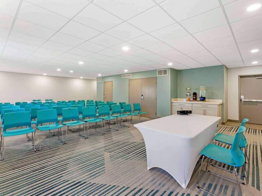 Uma sala de conferências com fileiras de cadeiras azul-turquesa, uma mesa branca e uma área de cozinha.