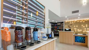 A modern coffee station with branded dispensers, a counter with a digital screen, and a view of the city through large windows. - Comfort Suites At Tucson Mall (Tucson)