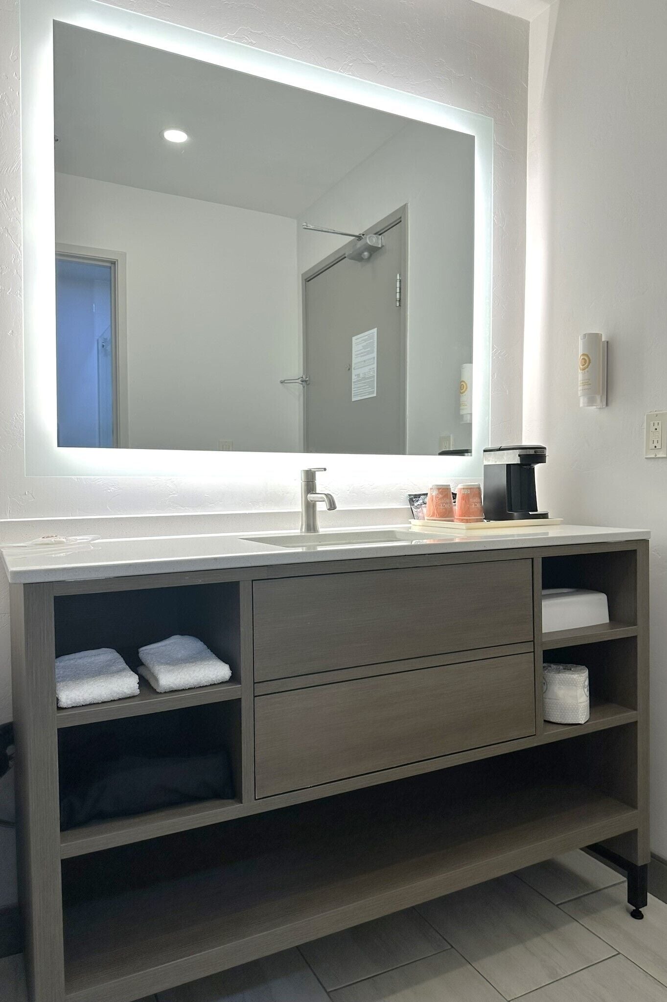 Ein modernes Badezimmer mit einem großen Spiegel, einem Waschbecken und einer Kaffeemaschine. Auf dem Waschtisch liegen Handtücher und ein Seifenspender.