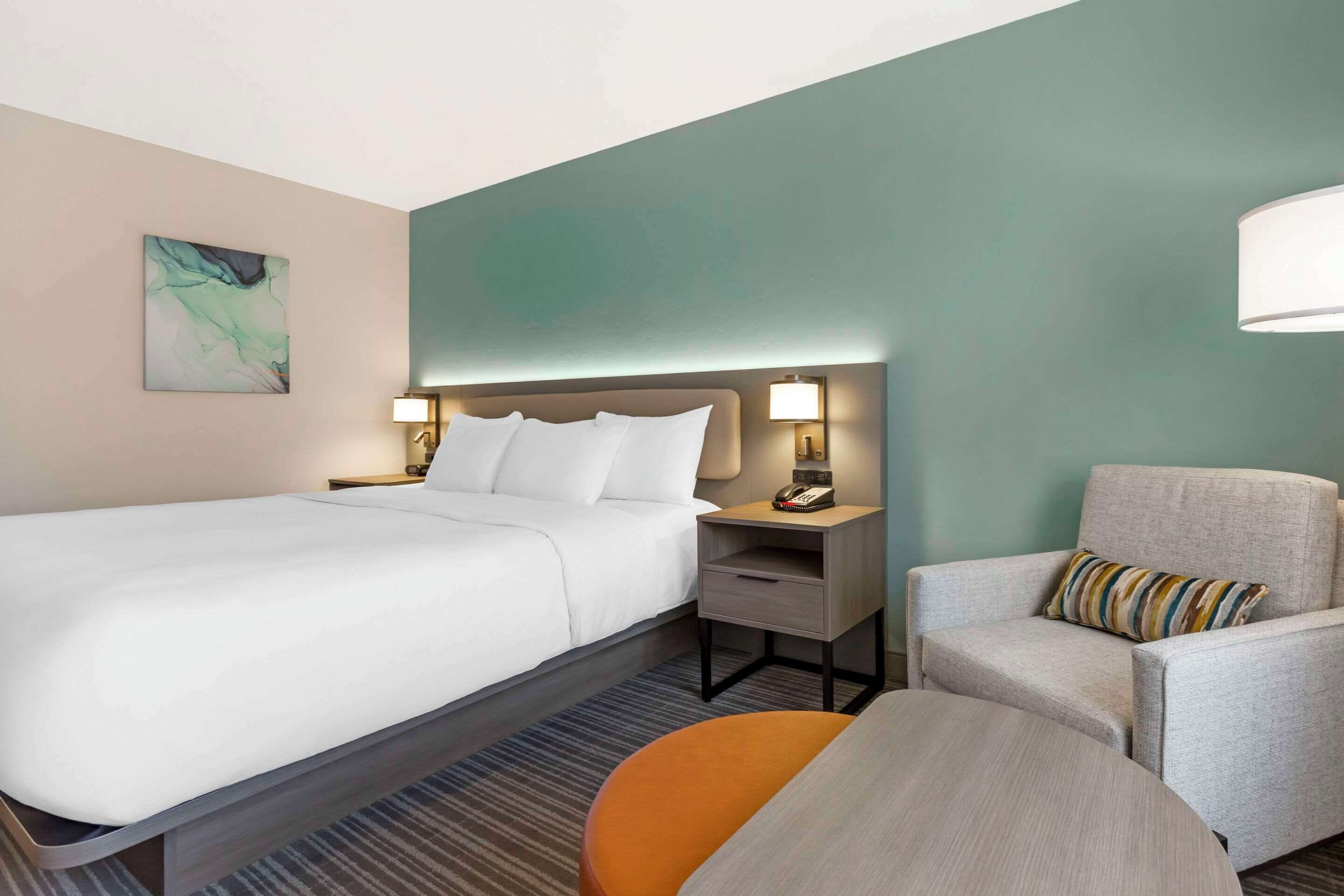 A hotel room with a large bed, a chair, a small table, and a nightstand with a lamp.