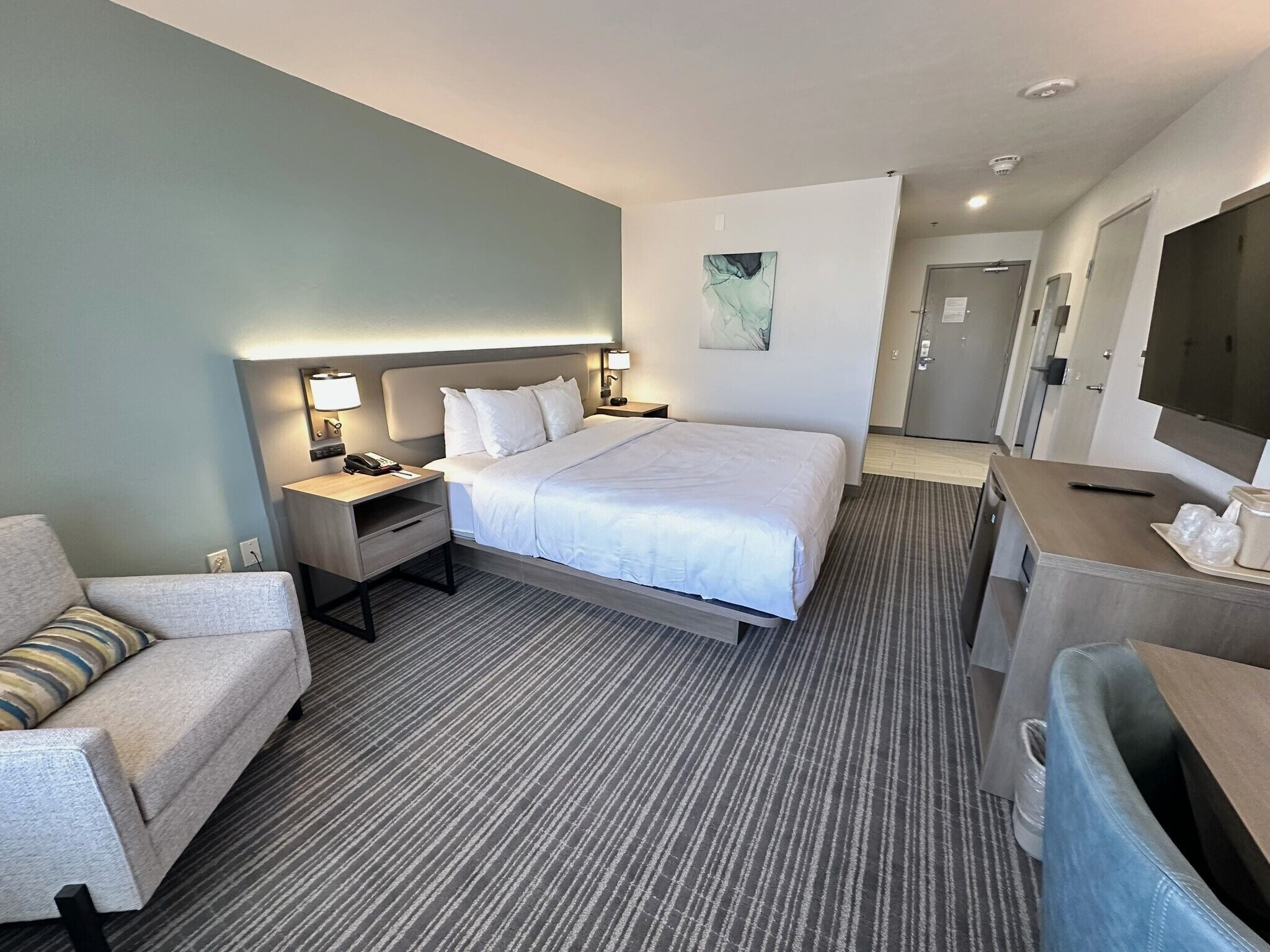 A hotel room with a large bed, a desk, a chair, a TV, and a bathroom visible in the background.