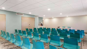 Uma sala de conferências com fileiras de cadeiras verde-água, uma mesa branca e uma parede bege.