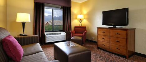 A hotel room with a sofa, ottoman, armchair, TV, dresser, and a window with curtains.
