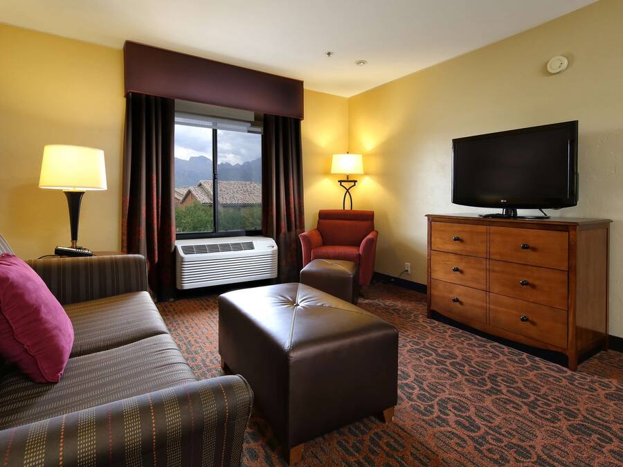 A hotel room with a sofa, ottoman, armchair, TV, dresser, and a window with curtains.