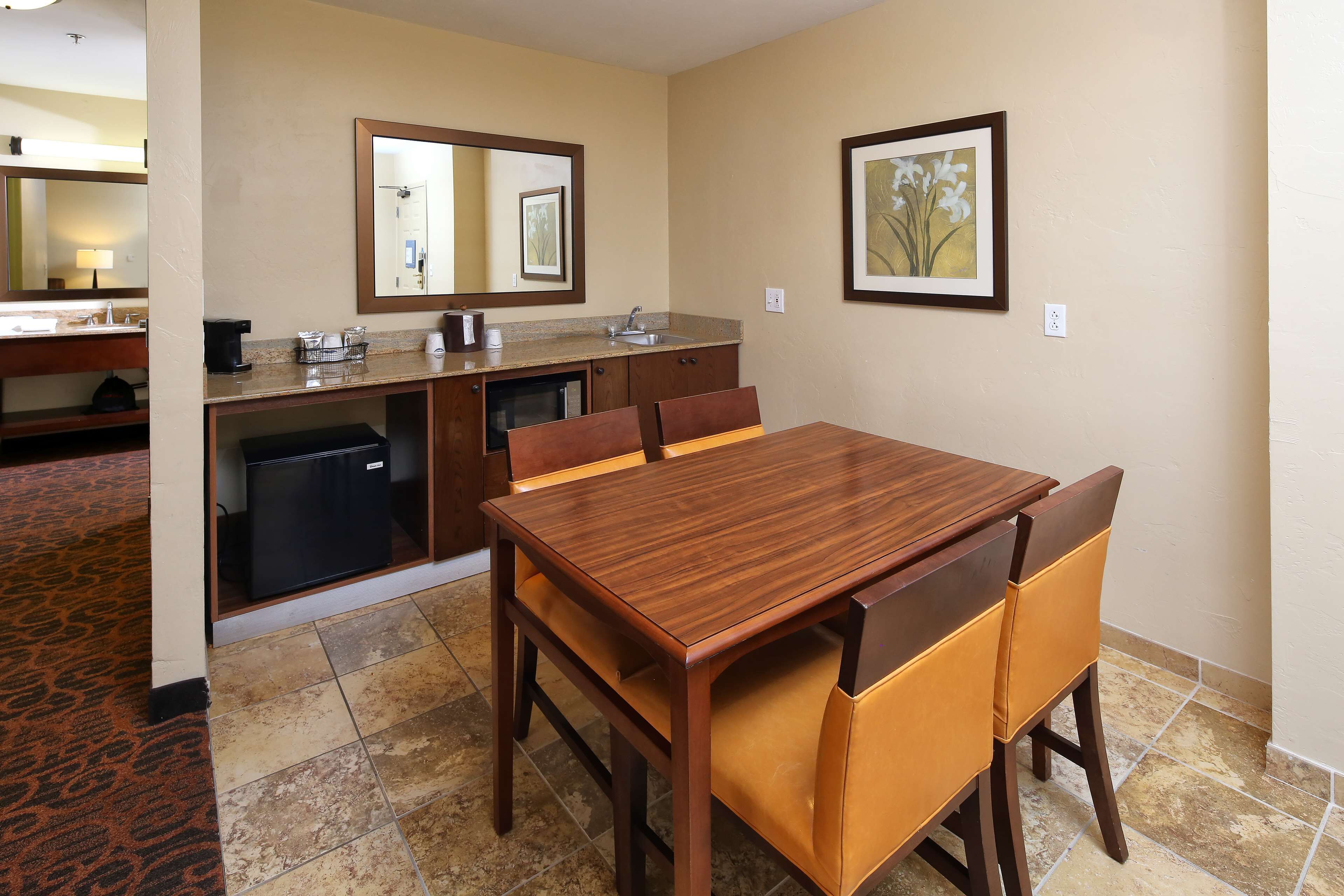 A hotel room with a wooden dining table, two chairs, a small kitchenette, a mirror, and a framed picture on the wall.