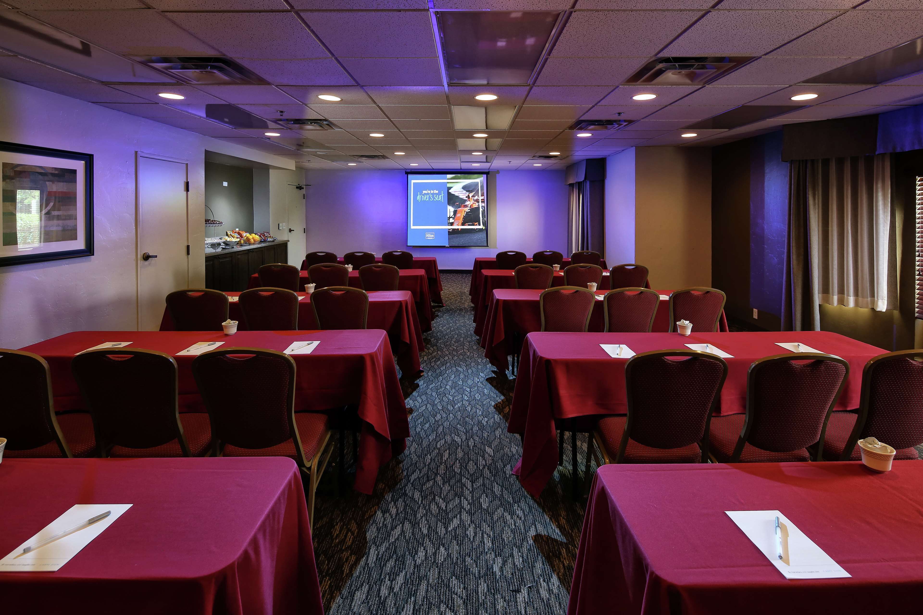 Une salle de conférence avec des rangées de tables et de chaises, un écran de projection et un espace buffet.
