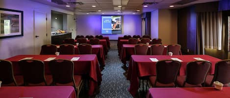 Une salle de conférence avec des rangées de tables et de chaises, un écran de projection et un espace buffet.